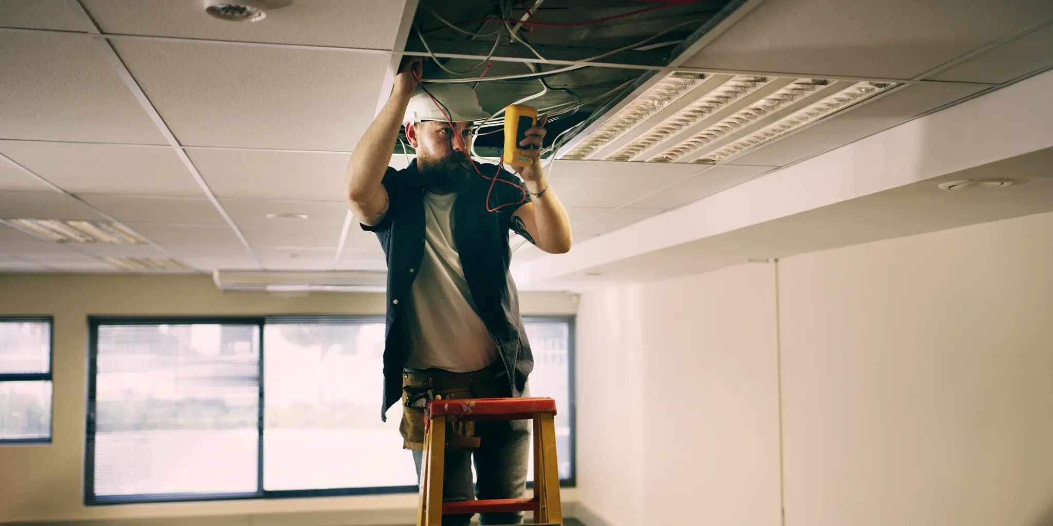 Elektriker auf Leiter prüft Kabel in abgehängter Decke eines Büros. Arbeitssicherheit, Installation, Technik.