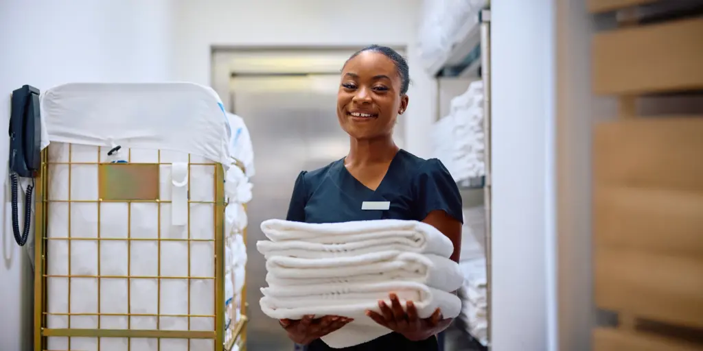 Frau in Uniform hält gestapelte Handtücher, steht lächelnd in einem Lagerraum mit Regalen voller Wäsche.
