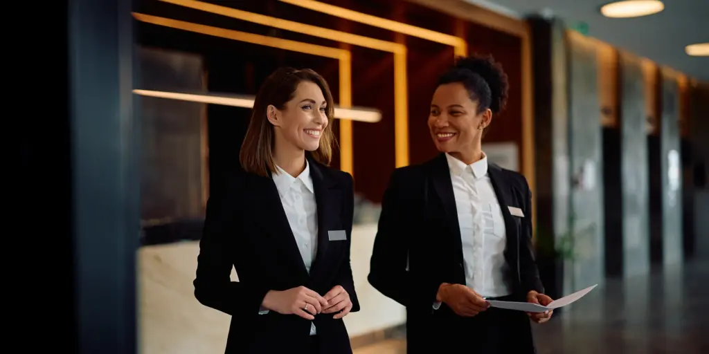 Zwei Frauen in Business-Kleidung lächeln und gehen in einem modernen Bürogebäude mit dunklen Holzverkleidungen.